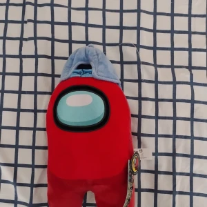 Among Us röd plushie med blå keps - Mjuk och färgglad Among Us plushie i rött med en ljusblå keps och broderat visir. Perfekt för fans av spelet eller som rolig inredningsdetalj. Officiell tagg sitter kvar på sidan.