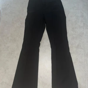 Svarta bootcut kostymbyxor från Amelie & Me - Snygga svarta kostymbyxor från Amelie & Me med bootcut-passform. Byxorna har hög midja och är tillverkade i en blandning av polyester och elastan för en bekväm känsla. Perfekta för dig som gillar klassisk stil med en modern twist.