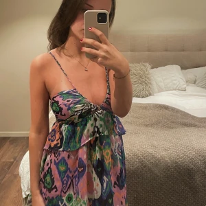 Färgglad kort klänning från Zara - Säljer en färgglad och somrig klänning från Zara i storlek XS. Säljs inte längre på Zara och dessutom oanvänd med lappen kvar!             Klänningen har smala axelband, v-ringning och ett mönster i lila, blått, grönt och rosa. Perfekt för varma dagar och har en luftig passform med volangdetalj vid midjan. 