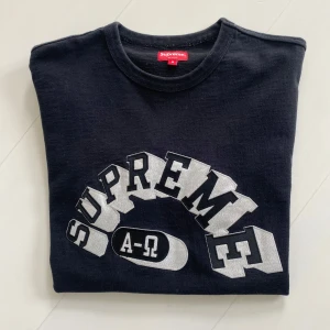 Svart Supreme t-shirt med tryck - Supreme t-shirt i svart med stort vitt tryck på bröstet där det står 'SUPREME' och 'A-Ω'. Klassisk rund hals och korta ärmar. Perfekt för dig som gillar streetwear och vill sticka ut med en ikonisk logga.