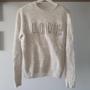 Grå stickad tröja med pärlbroderi - Mysig grå stickad tröja med texten 'LOVE' framtill, dekorerad med pärlor och glittriga stenar. Rund halsringning och långa ärmar. Perfekt för dig som gillar detaljer och vill ha en söt vardagströja. Knappt använd. 