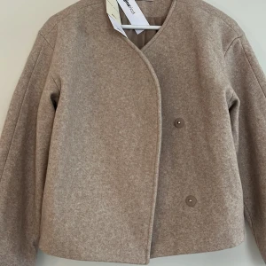 Beige jacka - Minimalistisk beige jacka  med asymmetrisk knäppning och två knappar framtill. Jackan har lång ärm och en rak, något boxig passform. Helt oanvänd med prislapp kvar.