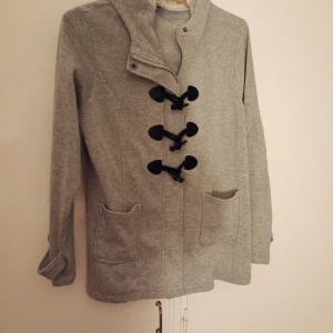 Grå kofta med huva - Säljer en grå kofta/tröja med huva och svarta knäppningar framtill. Jackan har två stora fickor och är i mjukt material, perfekt för kyliga dagar. Stilig och enkel design som passar till mycket.