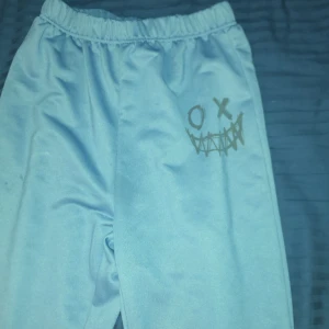 Ljusblå mjukisbyxor med tryck - Säljer ett par ljusblå mjukisbyxor med resår i midjan och ett tryck av ett smiley-ansikte på ena benet. Byxorna har en avslappnad passform och är perfekta för chill dagar. Mjukt material och enkel design.