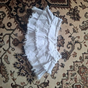 Vit volangkjol med fransar - Säljer en vit kortkjol med flera lager volanger och råa fransade kanter. Kjolen har en cool och avslappnad look, perfekt för dig som gillar att sticka ut. Materialet känns som bomull och kjolen har en mini-längd.