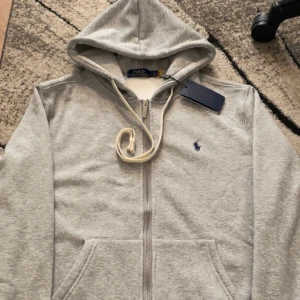 Polo Ralph lauren zip tröja - Säljer en klassisk grå hoodie från Polo Ralph Lauren med dragkedja och huva. Tröjan har en liten broderad logga i mörkblått på bröstet och vita snören vid huvan. Perfekt för en avslappnad stil.