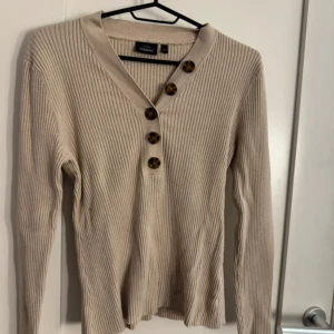 Beige ribbad långärmad tröja med knappar - Säljer en beige ribbad långärmad tröja från Weekday. Tröjan har v-ringning och fyra dekorativa knappar framtill. Perfekt för lager-på-lager och enkel att matcha med olika outfits.