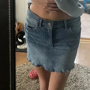Jeanskjol med rå kant - Säljer en klassisk blå jeanskjol med rå, fransig kant nertill. Kjolen har fem fickor och stängs med knapp och dragkedja framtill. Perfekt till vardags för en avslappnad look.