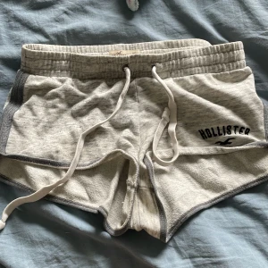 Grå mjukisshorts från Hollister - Superbekväma ljusgrå mjukisshorts från Hollister med mörkgrå kanter och vit dragsko i midjan. Shortsen har en broderad Hollister-logga på ena benet och är perfekta för en avslappnad stil.