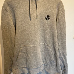 Grå hoodie från Jack & Jones - Säljer en stilren grå hoodie från Jack & Jones Premium med en svartvit patch på bröstet. Tröjan har huva med dragsko och en stor magficka framtill. Perfekt för en avslappnad look. Skriv om du undrar något 
