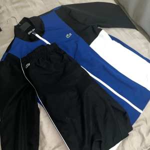 Blå och svart träningsdress från Lacoste - Säljer en snygg träningsdress från Lacoste i blått, svart och vitt. Setet består av en jacka med dragkedja och matchande byxor med resår i midjan. Perfekt för sport eller avslappnade dagar.