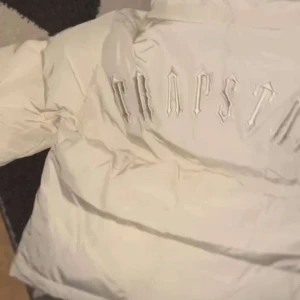 Vit pufferjacka från Trapstar - Säljer en vit pufferjacka från Trapstar med huva och dragkedja framtill. Jackan har Trapstar-logga broderad på ryggen och en rund logodetalj på bröstet. Perfekt för kyliga dagar.