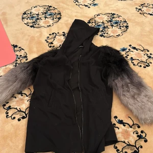 Svart jacka/kofta med gråa fuskpälsärmar och huva - Säljer en svart jacka med huva och dragkedja framtill. Ärmarna är täckta av långhårig grå och svart fuskpäls som ger en unik look. Perfekt för dig som vill sticka ut och hålla dig varm under kyliga dagar.