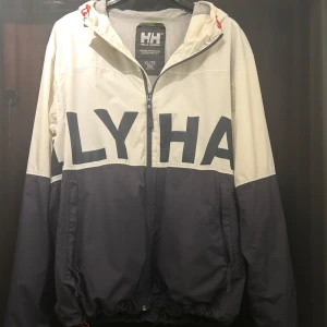 Helly Hansen vindjacka i blått och vitt - Säljer en snygg vindjacka från Helly Hansen i blått och vitt med stor logga över bröst och rygg. Jackan har huva, dragkedja och två fickor framtill. Perfekt för blåsiga dagar och enkel att matcha med det mesta.