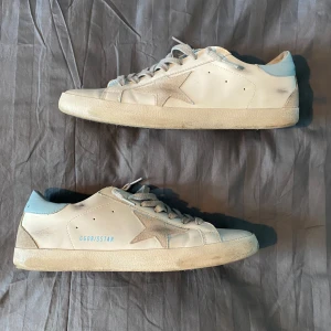 Golden Goose Superstar sneakers i vitt och ljusblått - Golden Goose Superstar sneakers i vitt skinn med ljusblå detaljer på häl och insida. Klassisk stjärndesign på sidan och snörning framtill. Platt sula och rund tå. Perfekta för en avslappnad och trendig look.