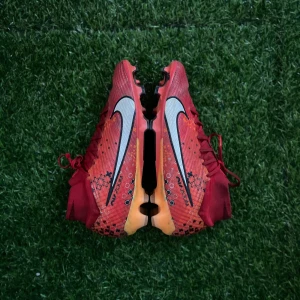Nike mercurial superfly elit MDS 007 - Säljer ett par röda Nike fotbollsskor med svart och vit swoosh på sidorna. Skorna har snörning och en platt sula med dobbar, perfekt för spel på gräs. Orange och svarta detaljer ger en sportig look. (Skick 8/10)
