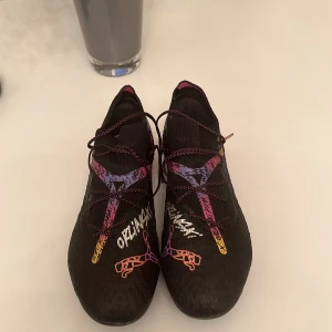 Limited edition oriliski puma future elit - Säljer ett par svarta Nike fotbollsskor med färgglada grafiska detaljer i lila, rosa och gult. Skorna har snörning och mönstrad sula med dobbar för bra grepp på planen. Snygg design med text och illustrationer på ovandelen.