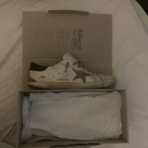 Golden Goose superstar med grå stjärna - Golden Goose sneakers i vitt skinn med grå stjärndetalj på sidan och grå snörning. Skon har en mörkblå häl och en platt sula. Dessa skor är perfekta inför sommaren. Skorna har sprickor i färgen fast det är bara att måla om dem. Allt og följer med har också kvitto som äktighets bevis 