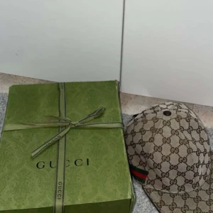 Gucci keps  - Säljer en beige keps från Gucci med klassiskt GG-monogram över hela kepsen och grönt/rött band på sidan. Justerbar rem baktill i skinn. Kepsen är i en stilren design och levereras med originalpåse och box. Priset ligger på 2300kr då den är äkta 