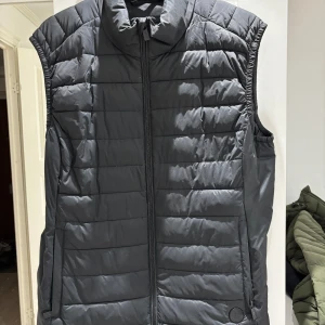 Svart dunväst från Snoot - Snygg svart dunväst från Snoot med quiltad design och dragkedja framtill. Västen har hög krage och två fickor med dragkedja. Perfekt för lager-på-lager och stilren look.