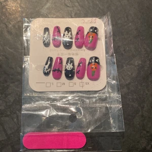 Halloween-inspirerade lösnaglar med motiv - Säljer ett set lösnaglar med halloweenmotiv. Naglarna är i svart och starkt rosa med detaljer som spindelnät, spöken och pumpor. Perfekt för dig som vill ha en unik och färgstark look till halloween eller temafest.