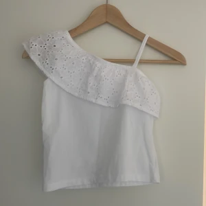 Vit one shoulder topp med volang - Säljer en vit one shoulder topp med broderad volangdetalj och smal axelrem. Toppen är ärmlös och har en romantisk känsla med sitt broderade mönster. Perfekt för varma dagar! Barnstorlek men typ XXS/XS, helt oanvänd för att jag inte gillar den 100% men det kostar att returnera. 