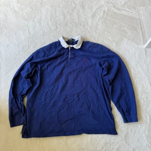 Blå långärmad pikétröja från Polo Ralph Lauren XXL - Säljer en klassisk blå långärmad pikétröja från Polo Ralph Lauren i 100% bomull. Tröjan har vit krage och en liten röd logga broderad på bröstet. Perfekt för en stilren och sportig look. En lite tjockare tröja så perfekt för sena sommarkvällar 