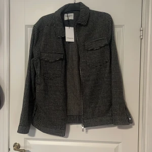 Svartvit rutig overshirt från These Glory Days - Säljer en stilren svartvit rutig overshirt från These Glory Days. Linne / Jackan har dragkedja framtill, klassisk krage och två bröstfickor. Perfekt lager-på-lager-plagg med normal passform.