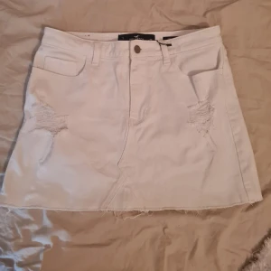Vit jeanskjol från Hollister - Säljer en vit jeanskjol från Hollister. Kjolen har slitna detaljer framtill, klassiska fem fickor och knappstängning. Säljer för att den inte passar mig. OBS Den har en fläck på baksidan! Den satt lite löst på mig som har xs i vanliga fall, men tror nog att den skulle se fantastisk ut på någon som har s eller m! Det står att den är high rise, men den faller ner på höften en del så skulle säga lite mitt emellan❤️