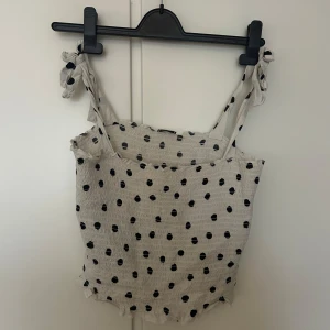 Vit prickig linnetopp från Pimkie - Supersöt vit linnetopp från Pimkie med svarta prickar och knytaxelband. Toppen har en smockad struktur och är perfekt för varma dagar. Säljer då den inte kommer till användning längre!