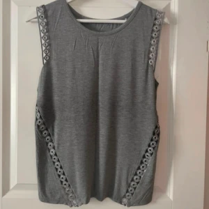 Grå ärmlös topp från Guess - Säljer en grå ärmlös topp från Guess med snygga detaljer längs axlar och sidor. Toppen har rund halsringning och är i mjukt material, perfekt för en stilren look. Aldrig använd.