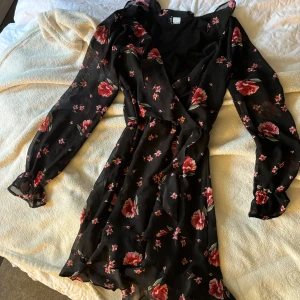 Svart blommig omlottklänning  - Supersöt svart omlottklänning från H&M med röda och rosa blommor. Klänningen har långa ärmar med lätt puff och volangdetaljer, samt v-ringad hals. Perfekt för vår och sommar!