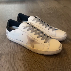 Skor från Golden Goose - Säljer ett par vita sneakers från Golden Goose med grå stjärndetalj på sidan och svarta hälpartier. Skorna har grå snörning och klassisk rund tå. Det är storlek 43. Ställ frågor vid fundering. 