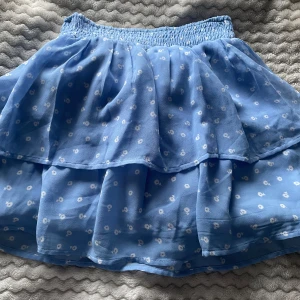Blå blommig volangkjol från Kappahl - Supersöt blå kjol från Kappahl med små vita blommor och dubbla volanger. Kjolen har resår i midjan för extra komfort och är perfekt för en somrig look. Är i storlek 134-140 men kan även passa 146-152. Den har en stretchig midja upp till ca 35cm. Skriv gärna om frågor eller om du är intresserad 😊