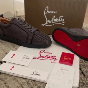 Grå sneakers  från Christian Louboutin - Säljer ett par gråa sneakers från Christian Louboutin med ikonisk röd sula och nitar på tån. Skorna har snörning och är tillverkade i skinn. Perfekta för dig som vill sticka ut med en lyxig och edgy stil.