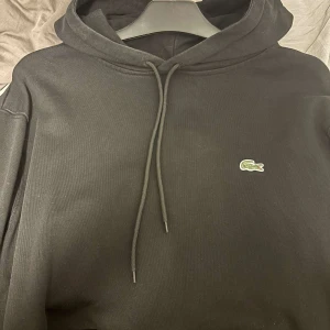 Svart hoodie från Lacoste - Säljer en svart hoodie från Lacoste med klassisk passform. Tröjan har huva med dragsko och den ikoniska krokodilloggan broderad på bröstet. Perfekt för en stilren och avslappnad look.