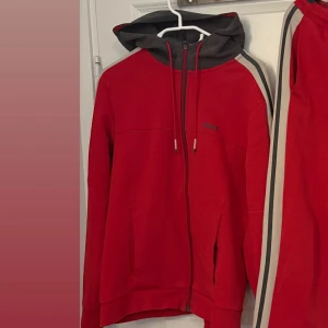 Röd hoodie från BOSS med huva - Röd hoodie från BOSS med svart huva och dragkedja framtill. Tröjan har snörning i huvan, diskret BOSS-logga på bröstet och beige-svarta ränder längs ärmarna. Perfekt för en sportig och avslappnad stil.