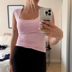 Ljusrosa ärmlös topp med spetskant - Supersöt ljusrosa topp med ärmlös design och fyrkantig ringning. Toppen har en fin vit spetskant vid urringningen som ger en extra detalj. Den är figurnära och gjord i mjukt bomullsmaterial, perfekt för en chill och feminin look. Inga defekter och använd fåtal gånger 