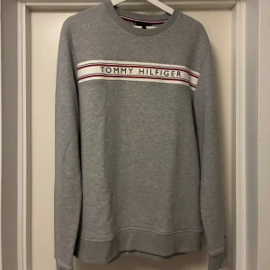  Tommy Hilfiger Sweatshirt - M - Snygg grå sweatshirt från Tommy Hilfiger i storlek M. Tröjan har rund hals, långa ärmar och ett broderat logoband med röd, vit och marinblå detalj över bröstet. Perfekt för en chill och stilren look.