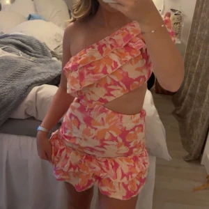 Blommig one shoulder kortklänning - Superfin kortklänning med blommigt mönster i rosa, orange och beige toner. Klänningen har one shoulder-design med volangdetaljer och en snygg cut-out i midjan. Perfekt för sommarens alla tillfällen och ger en riktigt trendig vibe. Aldrig använd utan endast testad! 🤩🌸