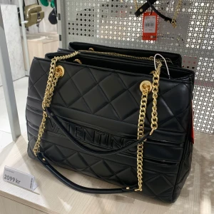 Svart väska från Valentino - (Äkta)Snygg svart axelväska från Valentino med quiltad design och guldfärgade kedjor. Väskan har ett stort VALENTINO-tryck framtill och detaljer i skinnimitation.köpte den för 2099kr och säljer den för 600kr 