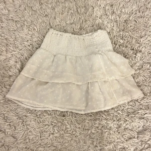 Vit volangkjol med smock i midjan - Supersöt vit kortkjol med volanger och smockad midja. Kjolen har diskreta prickar i tyget och är perfekt för en fräsch och somrig look. Lätt och luftig, passar perfekt till sneakers eller sandaler.