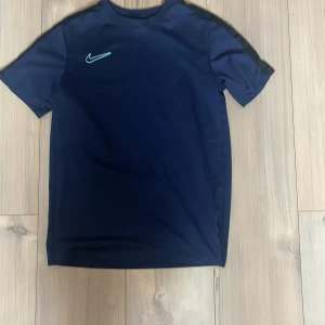 Mörkblå Nike Dri-Fit t-shirt 158-170 - Snygg mörkblå Nike Dri-Fit t-shirt med broderad ljusgrön swoosh på bröstet. Tillverkad i ett lätt och ventilerande polyestermaterial som passar perfekt för träning. Klassisk passform med korta ärmar och rund hals.