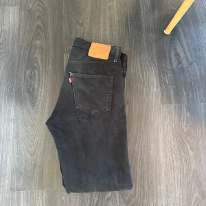 Svarta Levi's 501 jeans W32 L32 - Klassiska svarta Levi's 501 jeans med rak passform och femficksdesign. Jeansen har den ikoniska läderpatchen bak i midjan och är tillverkade i robust denim. Perfekta för dig som gillar en tidlös look och vill ha ett par jeans som funkar till allt.