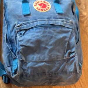 Blå Fjällräven Kånken ryggsäck - Klassisk blå Fjällräven Kånken ryggsäck med ikonisk rund logotyp framtill. Ryggsäcken har ett stort huvudfack, framficka med dragkedja och två sidofickor. Tillverkad i slitstark syntet med justerbara axelremmar och praktiskt handtag upptill. Lite snyggt slitage men även små hål i botten.