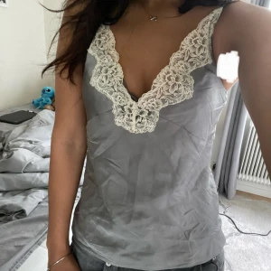 Grå satinlinnetopp med spets från Zara - Supersnygg grå linnetopp från Zara i satinliknande material med vit spetsdetalj längs v-ringningen. Topp med smala axelband och figurnära passform, perfekt att styla med jeans eller kjol. Ger en chic och trendig vibe! Kan alltid diskutera pris. Skriv för frågor och bilder🥰🥰