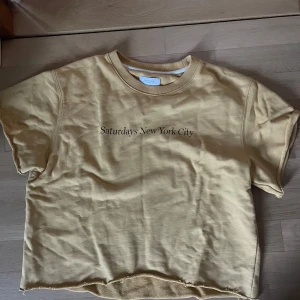 En beige/gul t-shirt från Saturdays New York City - Beige/gul t-shirt från Saturdays New York City med svart text på bröstet.