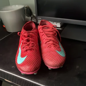 Nike Mercurial röda fotbollsskor - Säljer ett par Nike Mercurial fotbollsskor i rött med turkosa detaljer och snörning. Skorna har en smal, sportig siluett och är designade för konstgräs med dobbar undertill. Perfekta för dig som vill sticka ut på planen med stil.
