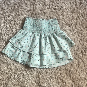 Ljusblå blommig volangkjol - Supersöt ljusblå kortkjol med blommigt mönster och bred smock i midjan. Kjolen har dubbla volanger som ger en flowy och lekfull känsla. Perfekt för vår och sommar när du vill ha en fräsch och romantisk look.