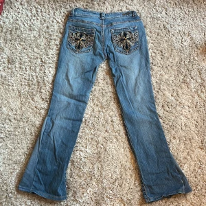 Blå lågmidjade bootcut jeans med broderade fickor - Snygga blå jeans med bootcut passform och unika broderade bakfickor i guld och svart. Klassisk femficksmodell i jeansmaterial, perfekt för dig som vill sticka ut me dom snygga fickorna. Väldigt lågmidjade 